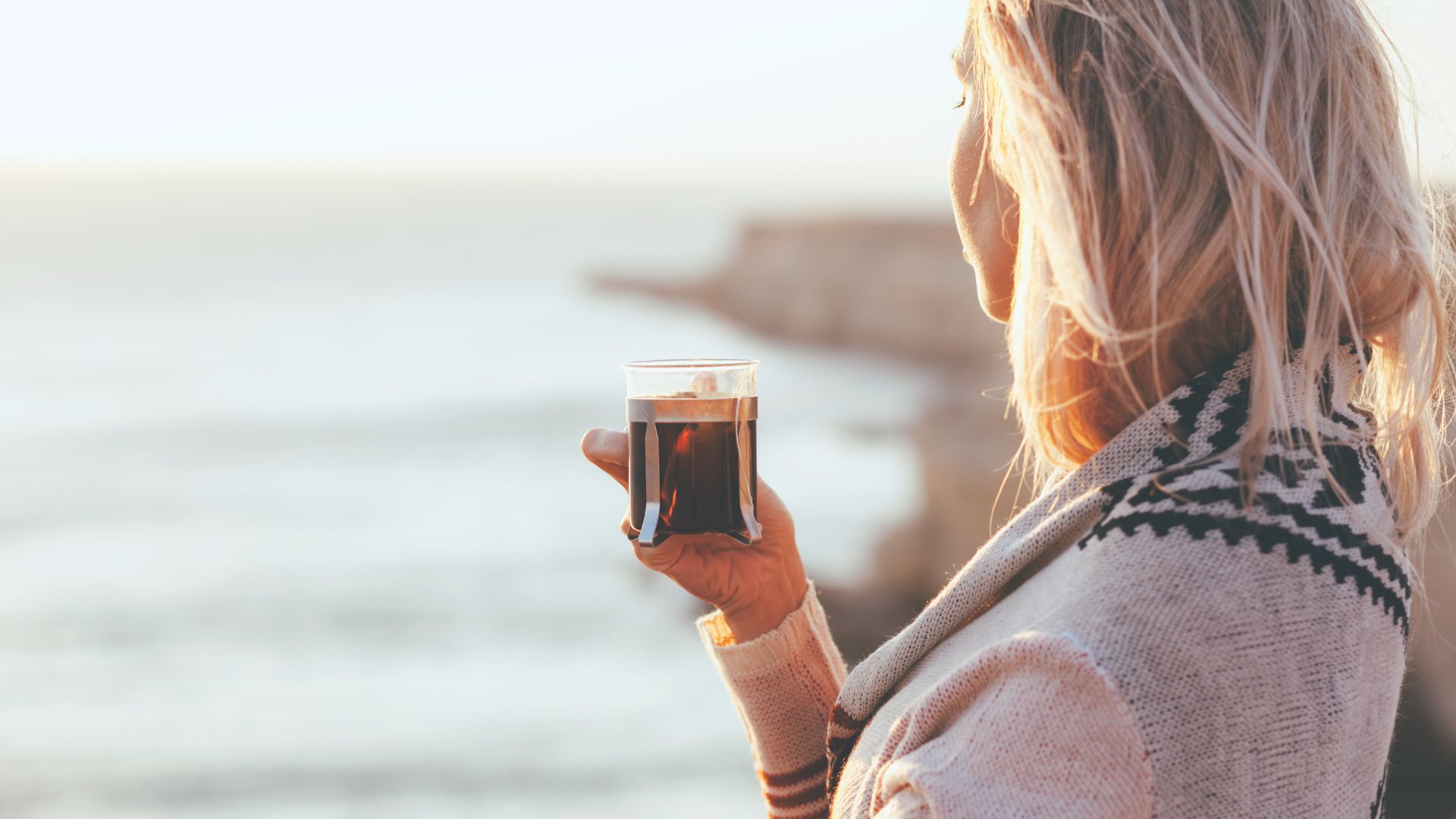 Vrouw van middelbare leeftijd kijkt uit over zee met kopje koffie in hand. Rustige reflectieve sfeer.