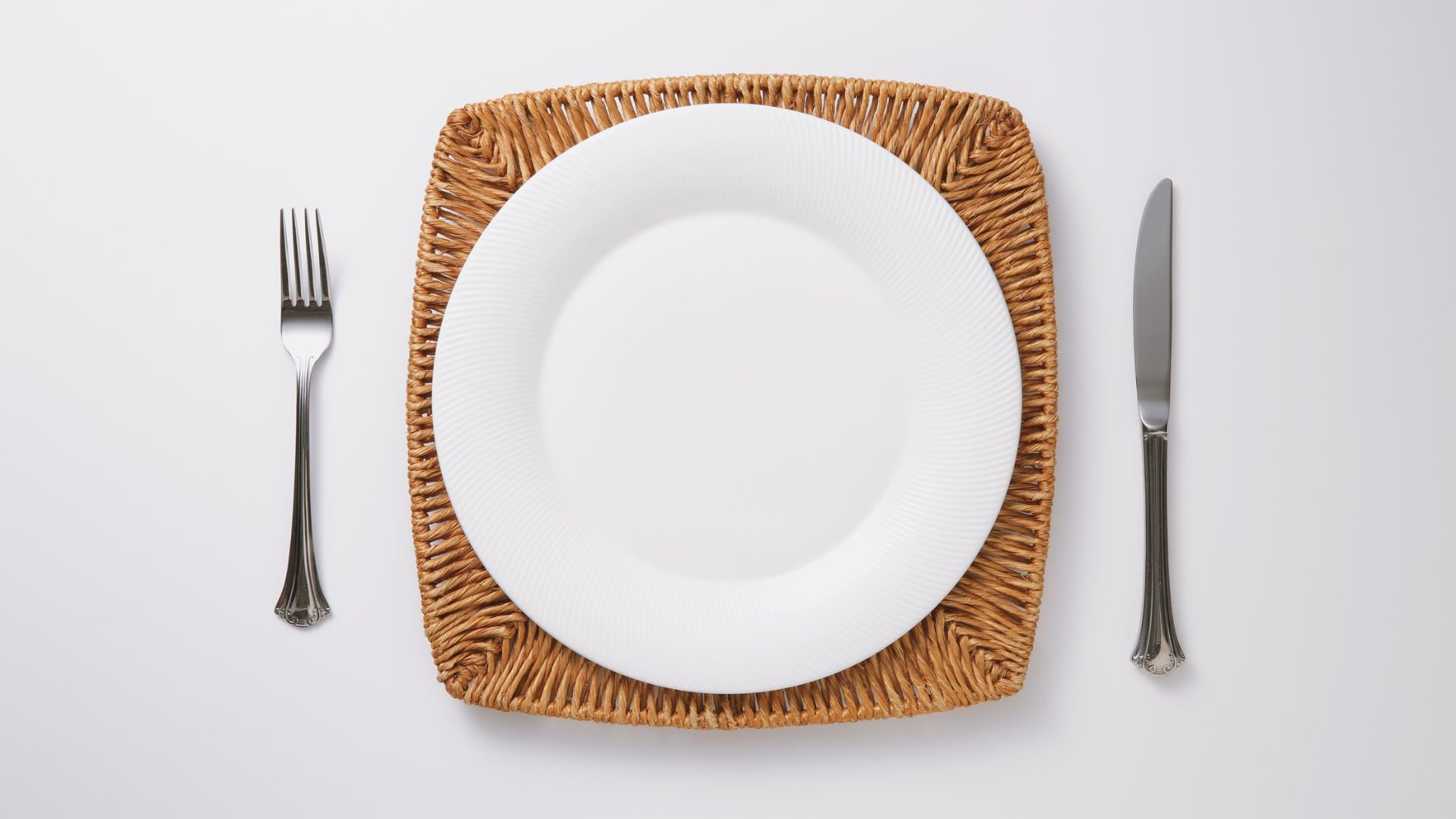 Afbeelding van een leeg bord op een gevlochten placemat met bestek — symboliseert stilte, ruimte of gemis in het eetpatroon.